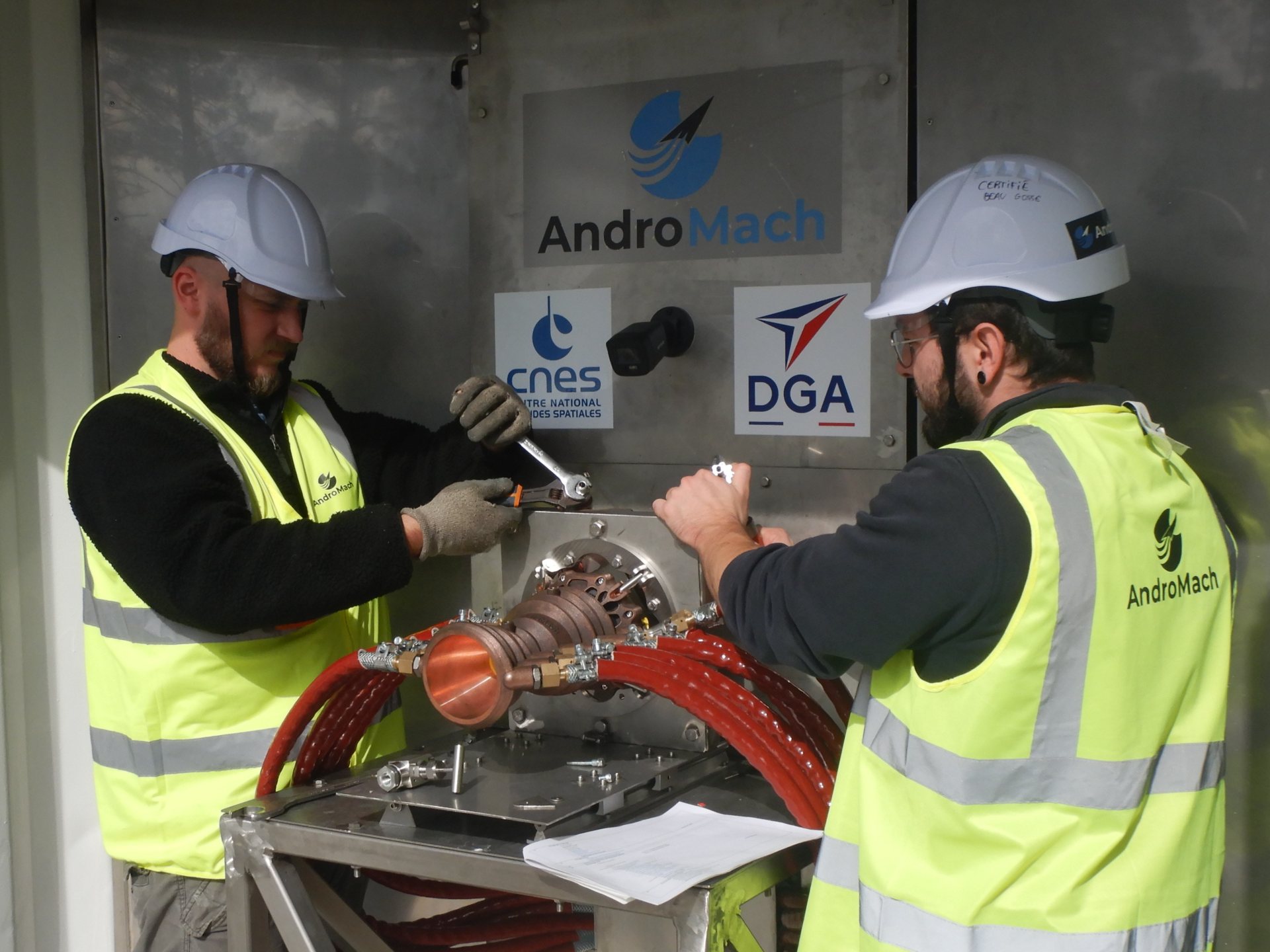 Two AndroMach engineers assembling the Banger engine by hand under the AndroMach, CNES and DGA panel